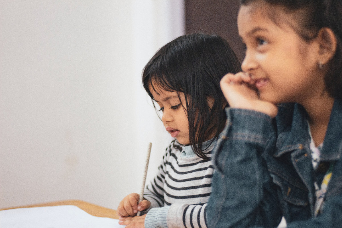 Two young girls with dark hair sit at a table together. One rests her chin on her hand, listening intently to someone out of frame. The other looks down at her paper and pencil.